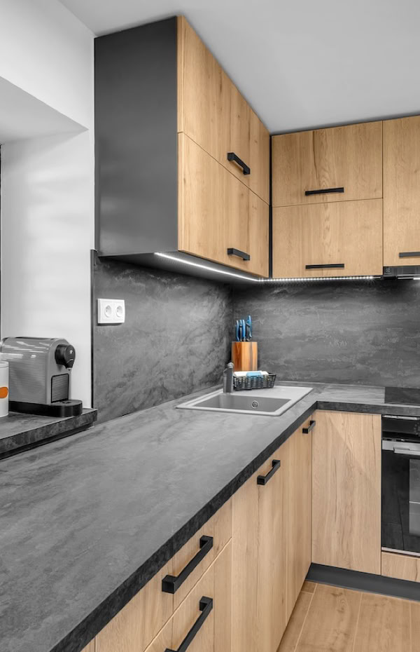 Modern kitchen with wooden cabinets, black countertops, a built-in oven, microwave, and various appliances on the counter near a window.