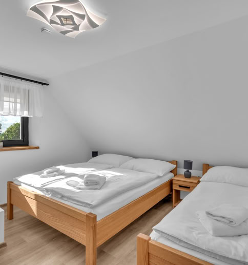 A bedroom with two wooden beds, each with white bedding and towels, a small bedside table between them, a window with sheer curtains, and a modern ceiling light fixture.