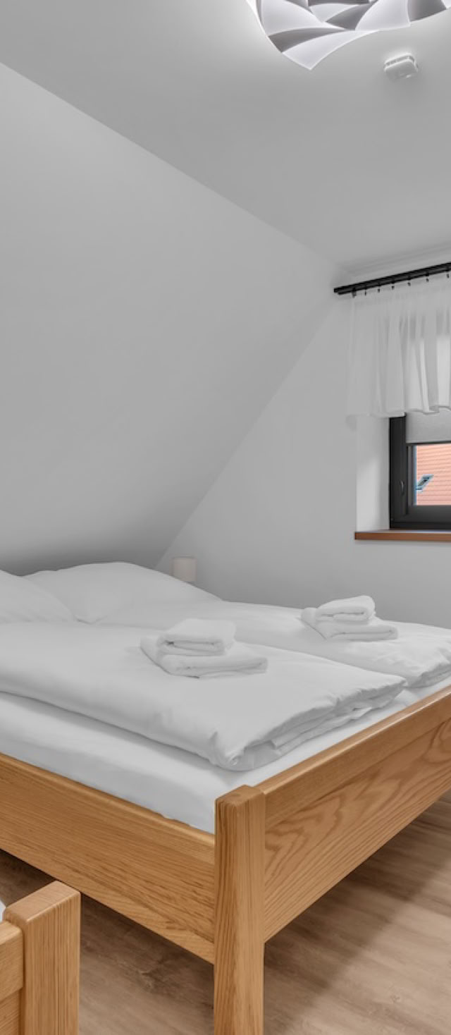 A small bedroom with two single beds, a wooden dresser with a television on top, a window with white curtains, and a nightstand between the beds.