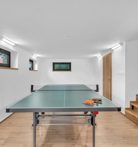A room with a ping pong table, wooden floor, white walls, small windows, and a staircase.