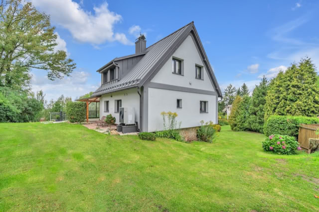 A two-story house with a gray roof and white walls, surrounded by a green lawn and trees under a blue sky with clouds.