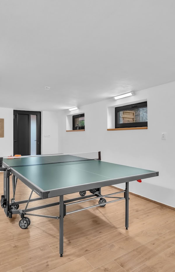 A room with a ping pong table on a wooden floor, white walls, and small windows near the ceiling.