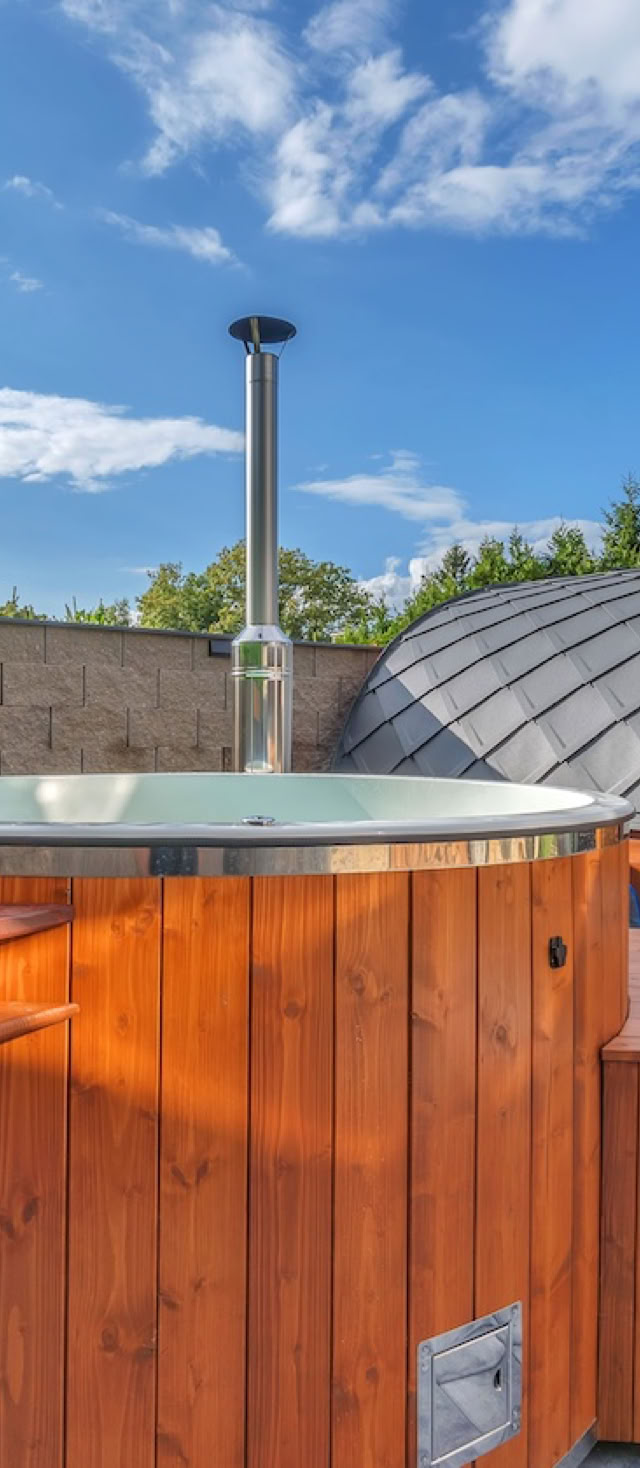 Wooden hot tub with steps in an outdoor setting, adjacent to a barrel-shaped sauna, with a stone wall and greenery in the background under a blue sky.