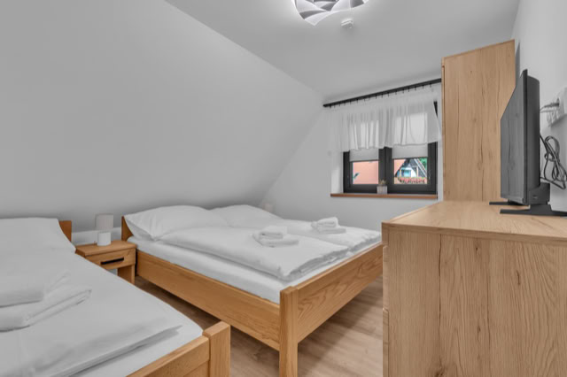 A small bedroom with two single beds, a wooden dresser with a television on top, a window with white curtains, and a nightstand between the beds.