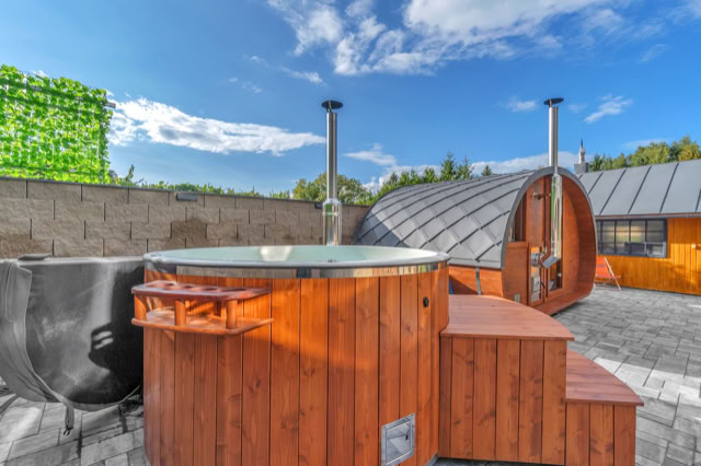 Wooden hot tub with steps in an outdoor setting, adjacent to a barrel-shaped sauna, with a stone wall and greenery in the background under a blue sky.