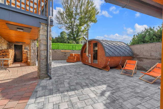 A courtyard with a wooden pod structure, two orange deck chairs, stone walls, and a tree in the background under a blue sky.